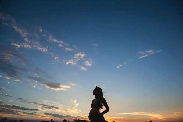 A pregnant woman stands before a sunset with her hands on her belly.