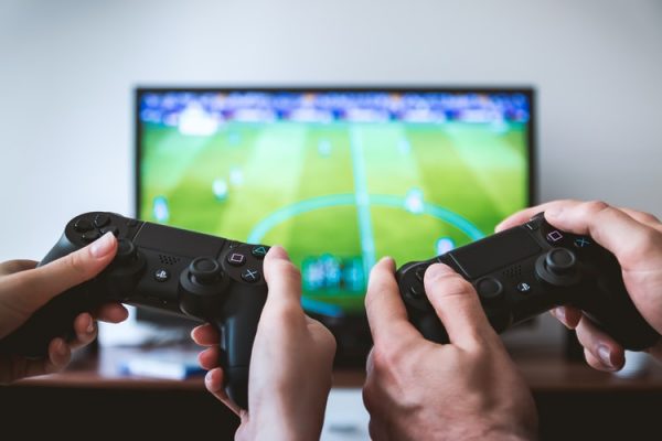 Two pairs of hands hold up video game controllers in front of a TV.