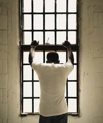 A person in a white shirt stands in front of a tall window with their back to the viewer and arms resting on the window.